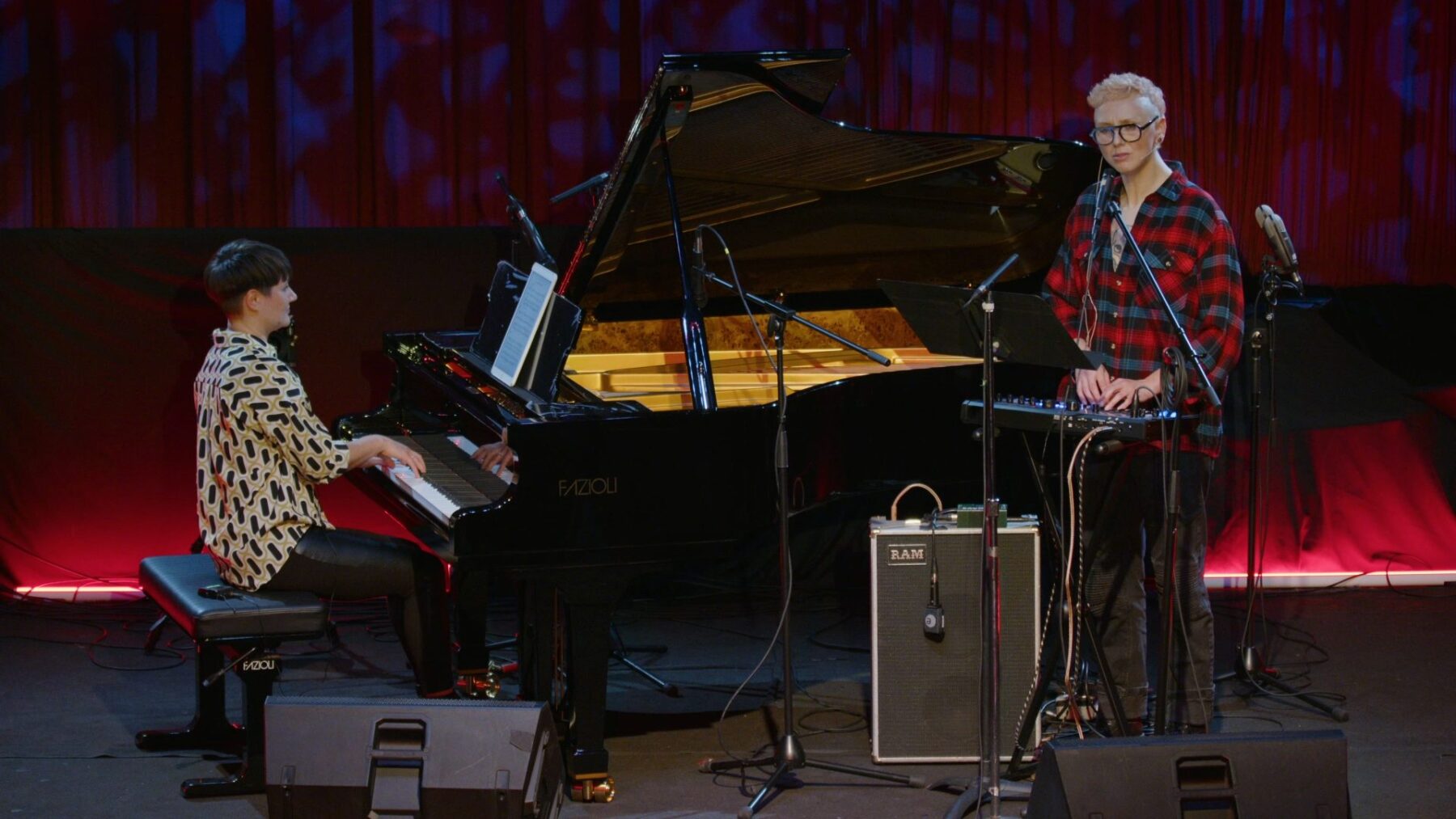 Madeline Hildebrand sitting at the piano and Sarah Jo Kirsch standing at a microphone in front of a red background.