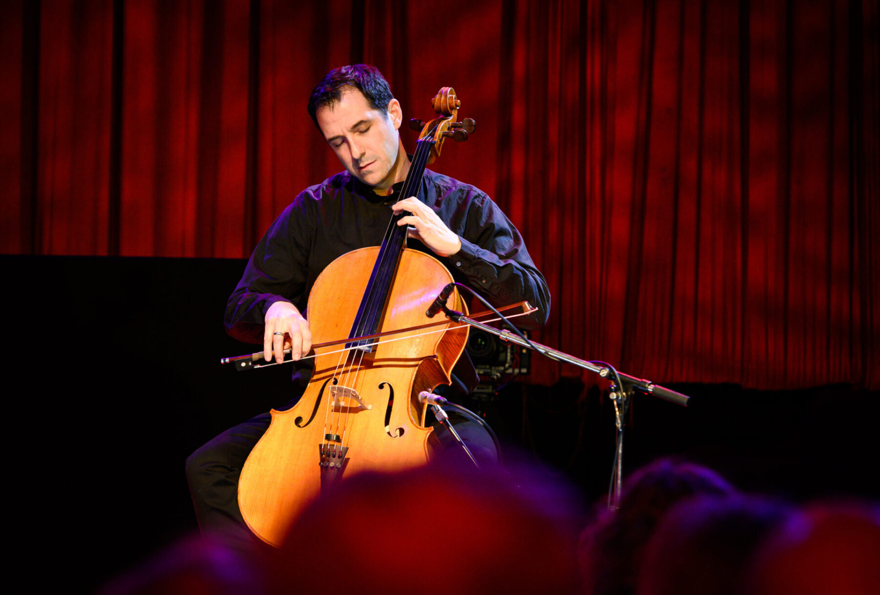 Ariel Barnes in a black outfit, playing the cello in front of a red background.