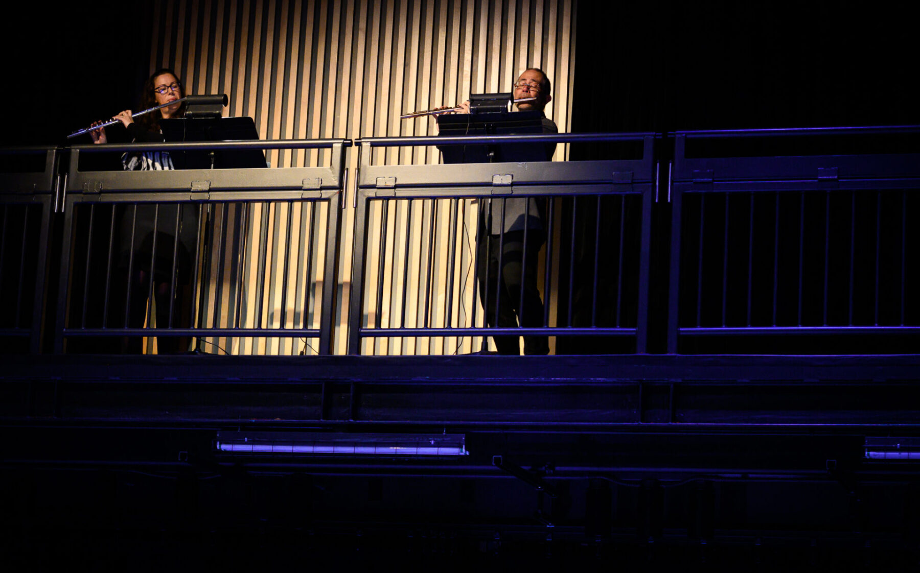 Jennifer Butler and Mark Takeshi McGregor playing flutes while spotlit on a wood frame backgroun.