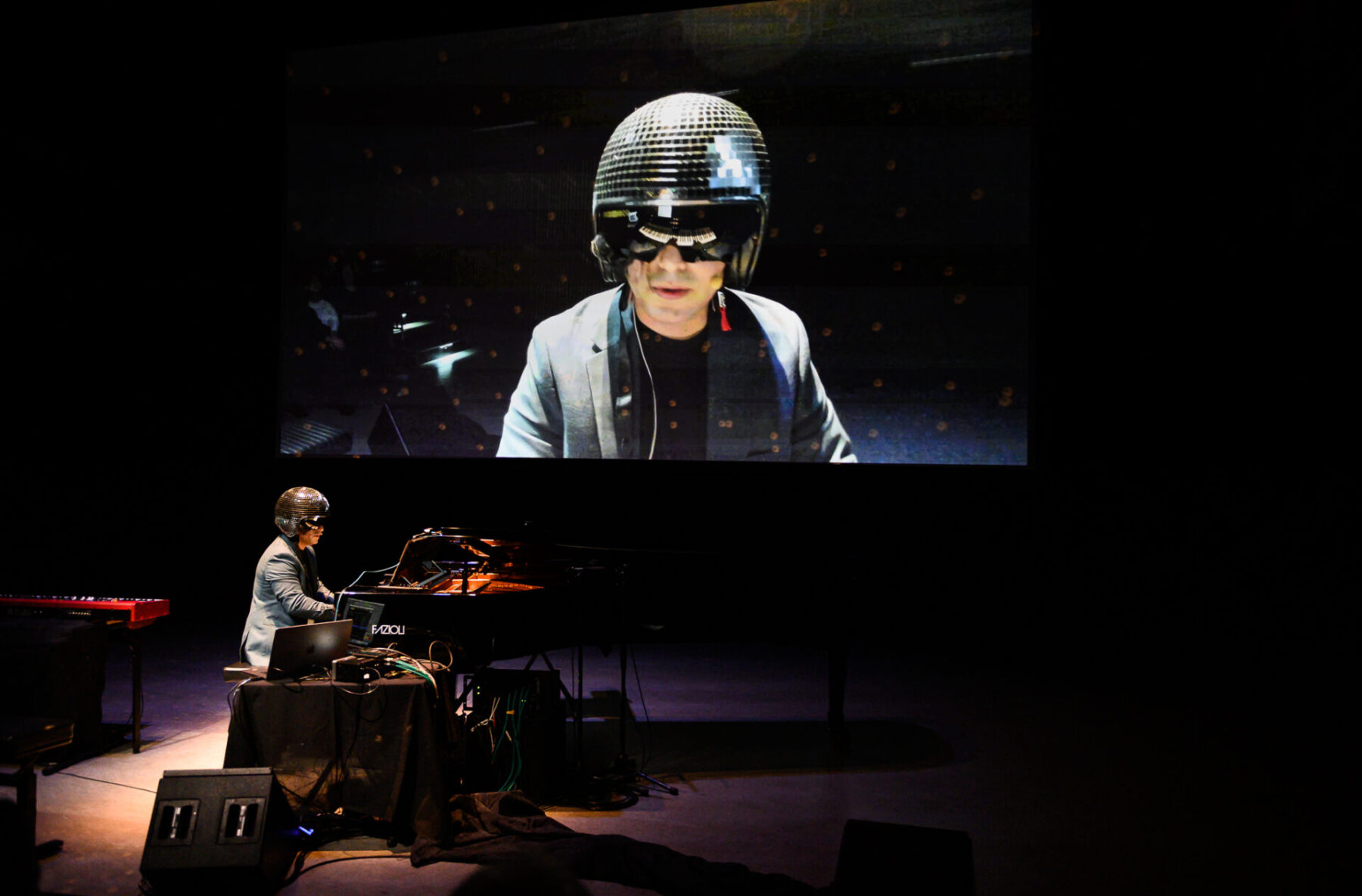 Zubin Kanga performing at a piano in front of screen showing him wearing a disco ball helmet.