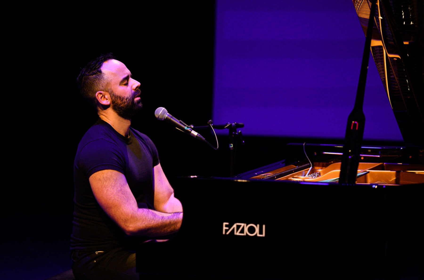 Adam Tendler in a black t-shirt sitting at the piano while speaking into a microphone.