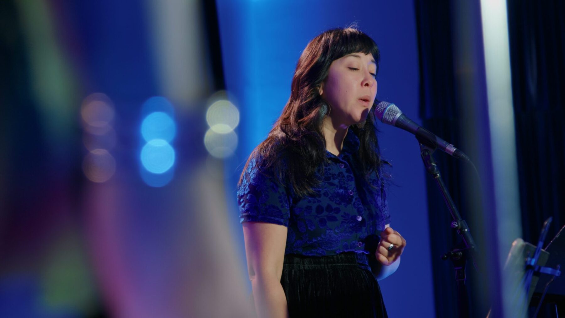 Robyn Jacob wearing a blue dress, singing into a microphone with a sparkly yet blurry detail around her.