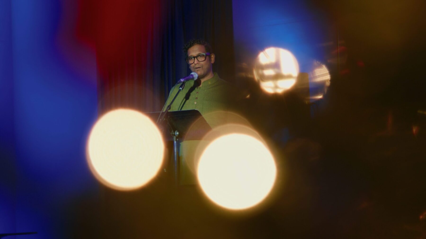 Asitha Tennekoon wearing a green shirt, singing into a microphone with a sparkly yet blurry detail around him.